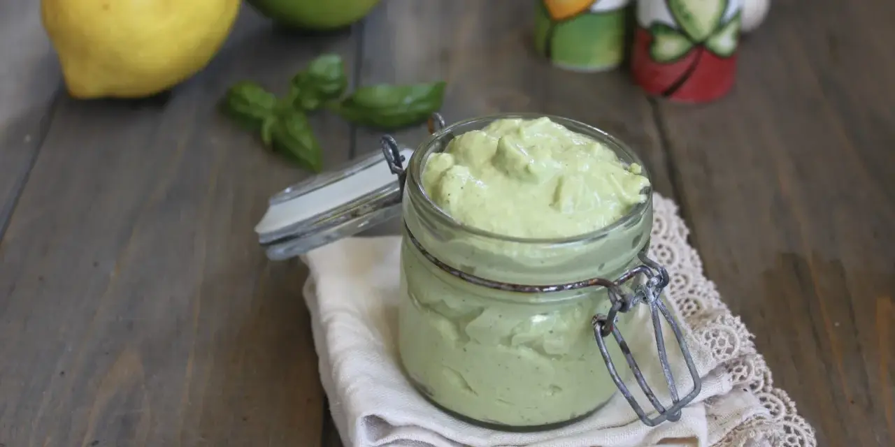 Un vasetto di cremoso pesto al limone, con limoni e basilico sullo sfondo.