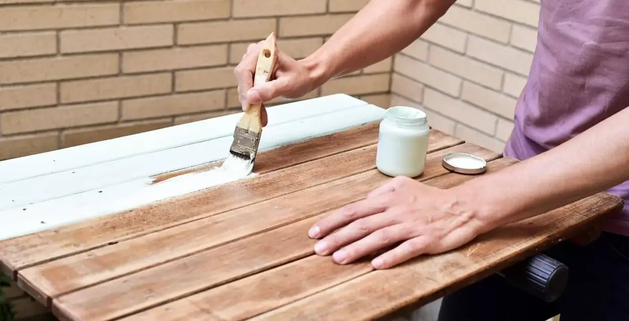 Persona pinta madera de blanco con brocha. Un frasco de pintura blanca y su tapa están cerca.
