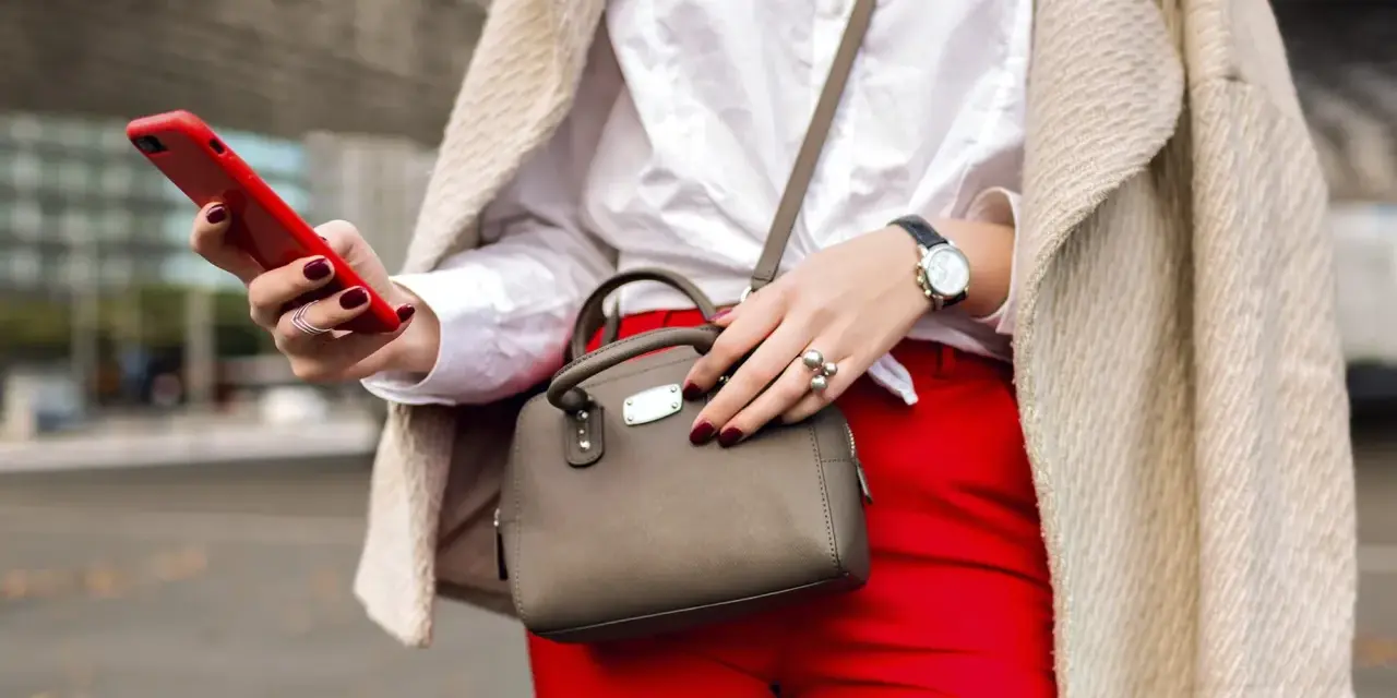 Une femme élégante, avec un manteau beige, un chemisier blanc et un pantalon rouge vif, tient un téléphone rouge et un petit sac gris. Quel entoilage pour sac choisir pour un tel look ?