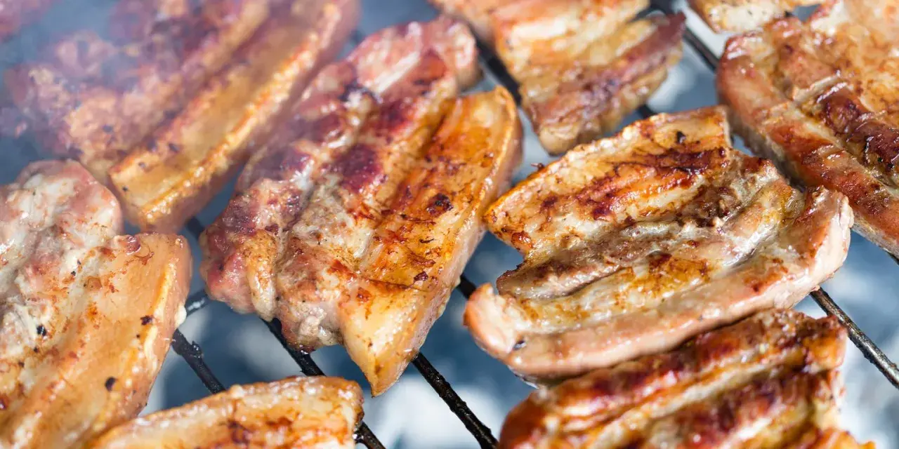 Kawałki soczystego boczku surowego pieką się na grillu, otoczone dymem.
