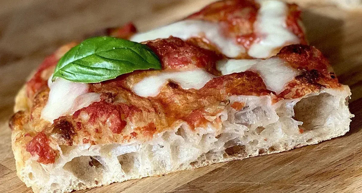 Fetta di pizza fatta in casa teglia, con crosta alveolata, pomodoro, mozzarella filante e una foglia di basilico fresco.