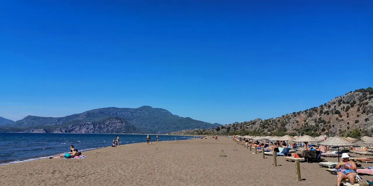 Słoneczna plaża w Sarigerme, z turkusowym morzem, górami w tle i rzędami leżaków pod słomianymi parasolami.