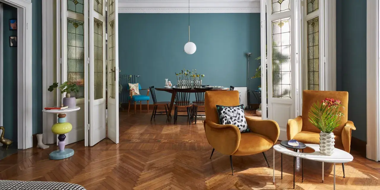 Salotto con un mix di arredamento moderno e antico: poltrone ocra, tavolino colorato, porte a vetri d'epoca e un tavolo da pranzo in stile classico.