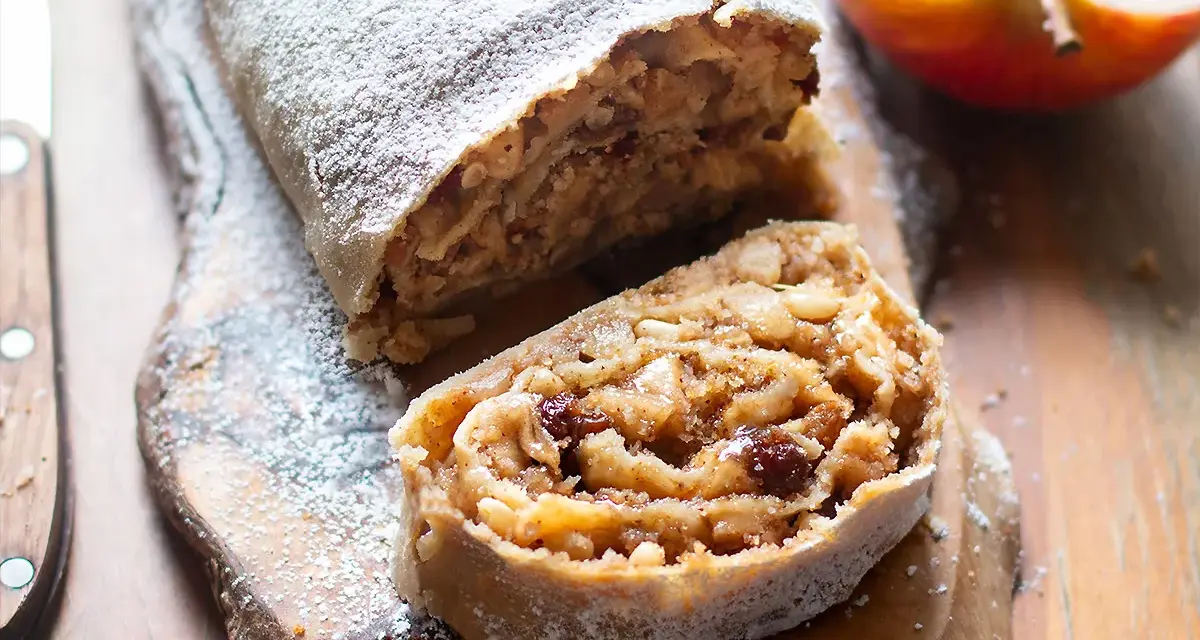 Un delizioso strudel di mele, cosparso di zucchero a velo, con un morso invitante che rivela il ripieno di mele, uvetta e pinoli.