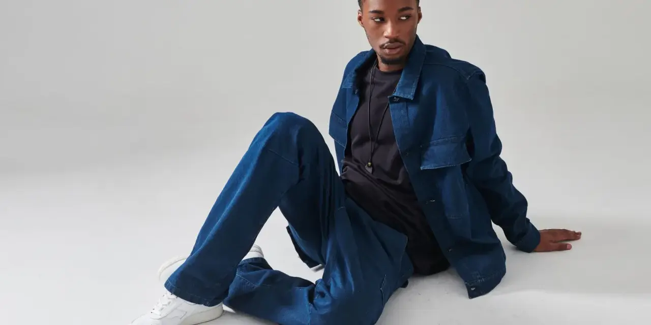 Giovane uomo con completo in materiale jeans, seduto a terra, con giacca e pantaloni oversize.