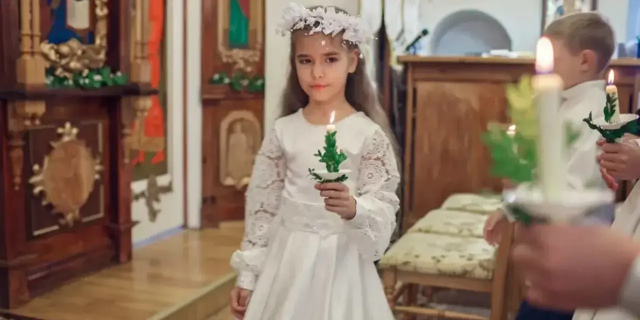 Dziewczynka w białej sukni i wianku trzyma świecę. To ważny dzień, w której klasie komunia.