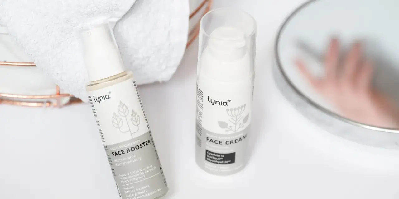 Białe opakowania kosmetyków Lynia: Face Booster i Face Cream, z widocznym składnikiem papaina. Obok ręczniki i lustro.