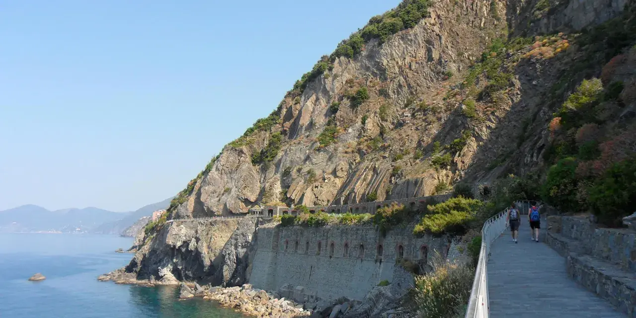 Wanderer auf dem Sentiero Azzurro entlang der felsigen Küste Liguriens. Das Meer glitzert türkisblau neben dem steilen Hang.