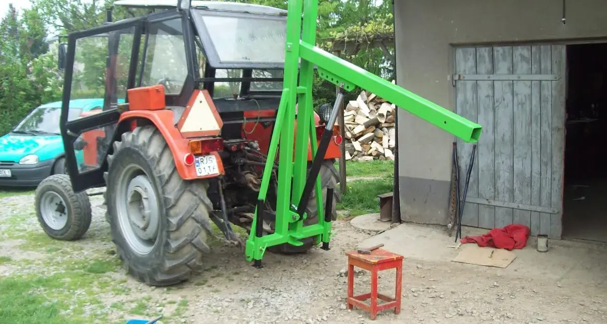 Czerwony traktor z zamontowanym zielonym ładowaczem TUZ przed budynkiem gospodarczym.