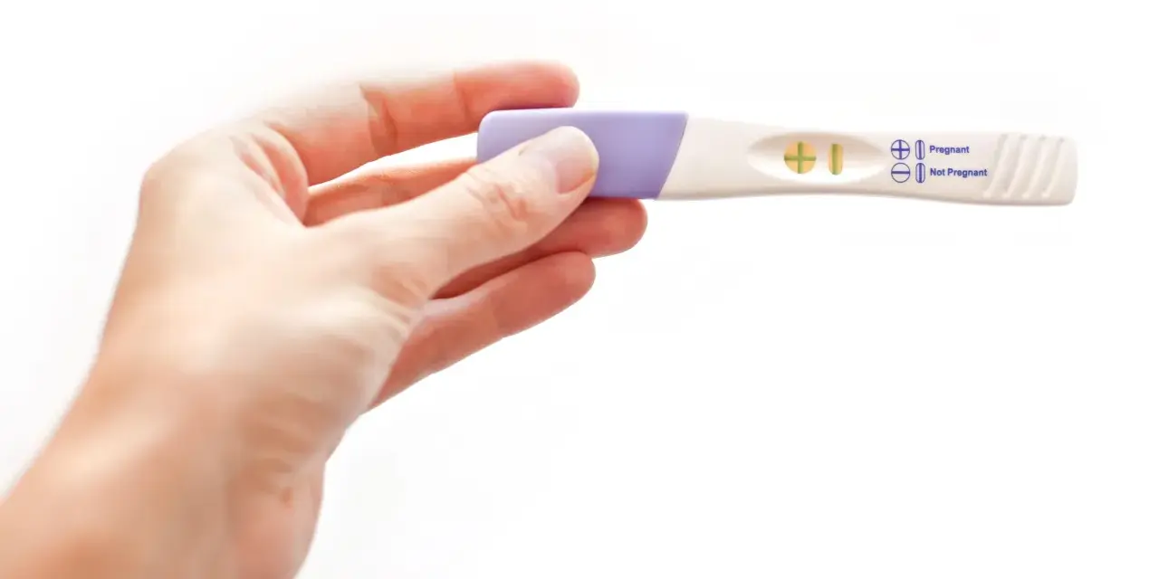 Dłoń trzyma test ciążowy z wynikiem pozytywnym. W okienku widoczny jest symbol plusa, oznaczający ciążę.