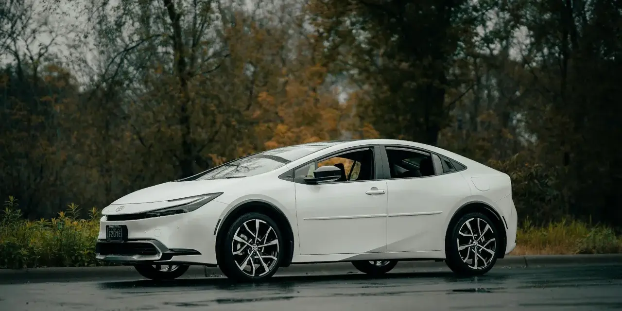 Biała Toyota Prius zaparkowana na mokrej drodze, z drzewami w tle.