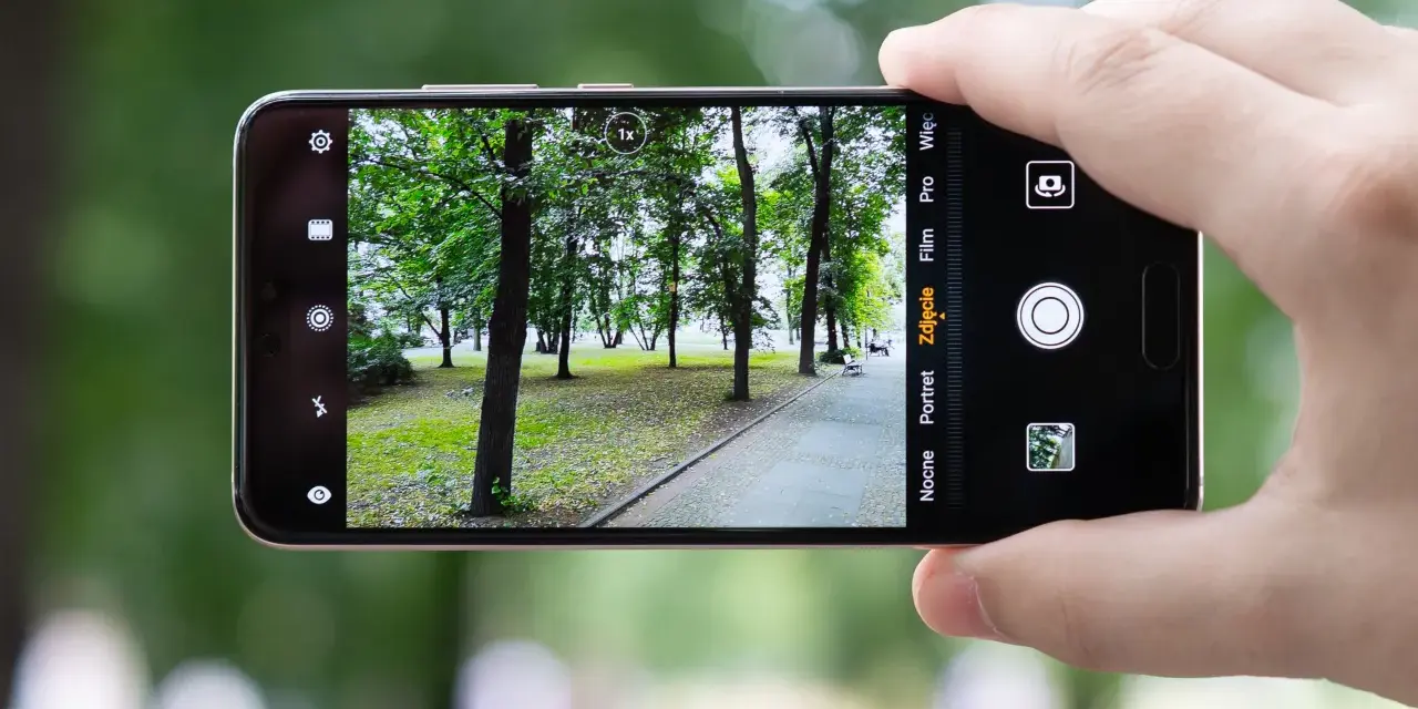 Dłoń trzyma smartfon Huawei P20, aparat kadruje parkowy krajobraz z alejką i drzewami.