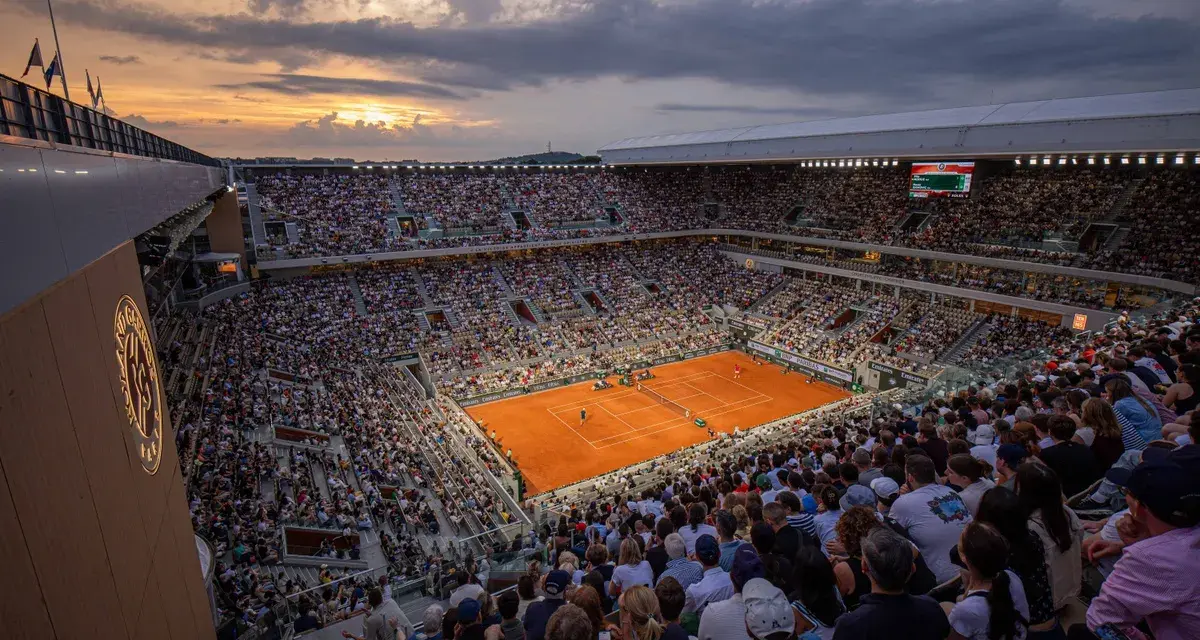 Widok na pełny stadion podczas meczu na kortach ziemnych. Zachodzące słońce oświetla trybuny pełne widzów, tworząc atmosferę emocjonującego **Roland Garros live**.