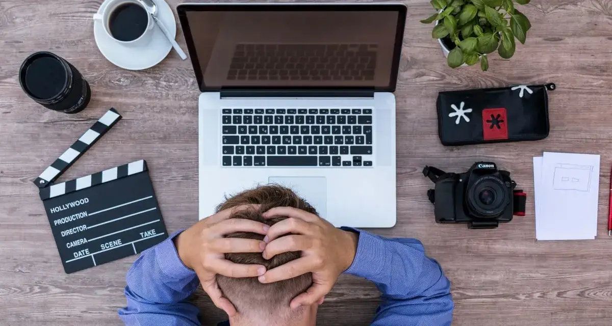 Frustracja twórcy filmowego. Na drewnianym biurku leży laptop, aparat, klaps, kawa i roślina.