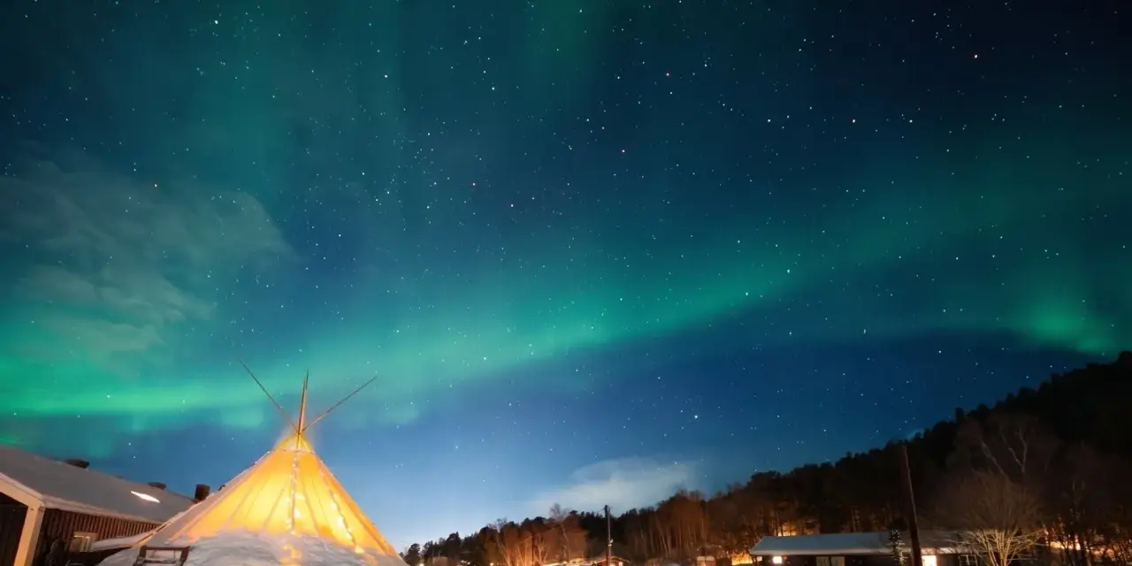 Nocne niebo nad ośnieżoną wioską, gdzie nie zachodzi słońce. Tańcząca zorza polarna nad oświetlonym tipi i domkami.