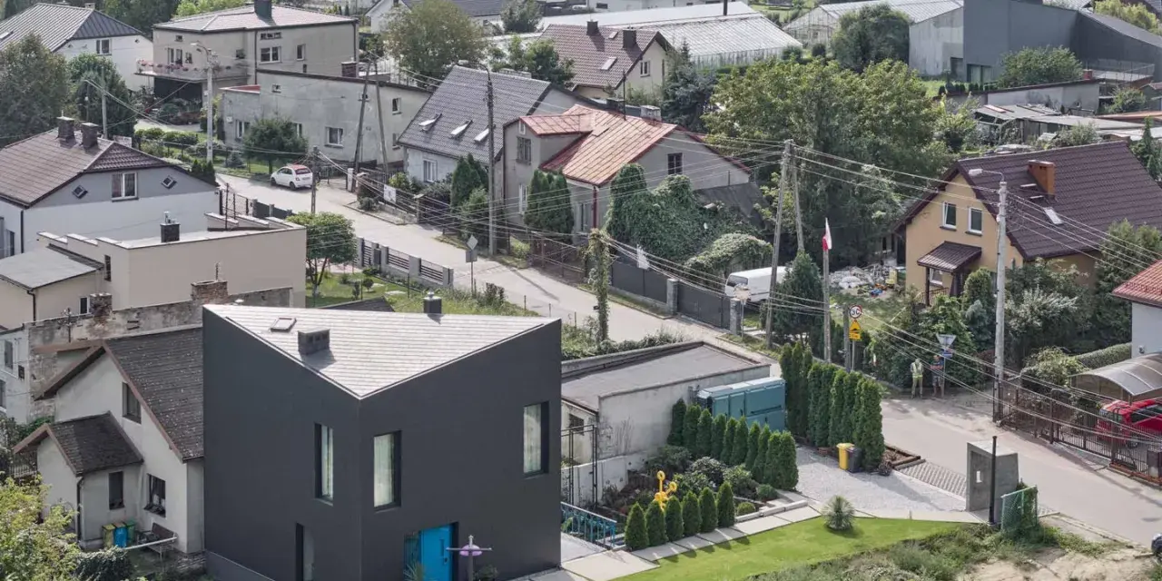 Nowoczesny, czarny dom o trójkątnym kształcie wyróżnia się na tle tradycyjnych terenów mieszkaniowych b. Otoczony zielenią, z niebieskimi drzwiami.