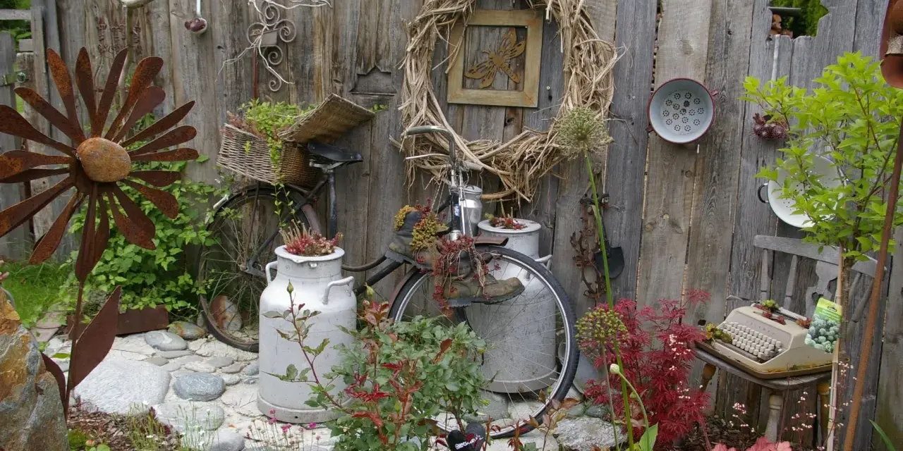 Kreative Gartendeko Ideen: altes Fahrrad mit Blumen, Milchkannen, Rostblume und Schreibmaschine vor Holzzaun.