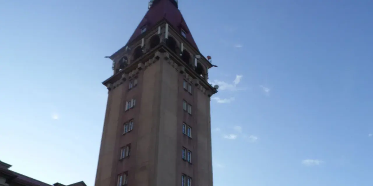 Wysoka wieża z czerwonym dachem, przypominająca dom rybaka, na tle błękitnego nieba.