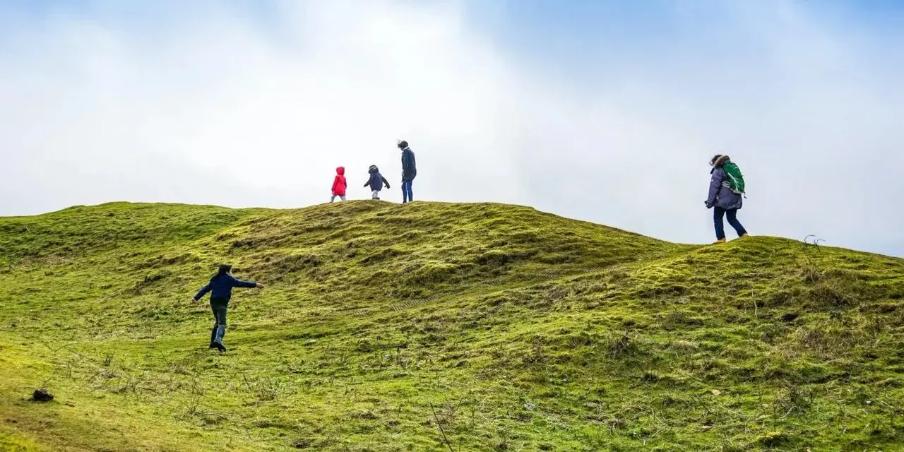 Famiglia esplora sentieri per bambini su una collina verdeggiante sotto un cielo azzurro e nuvoloso.