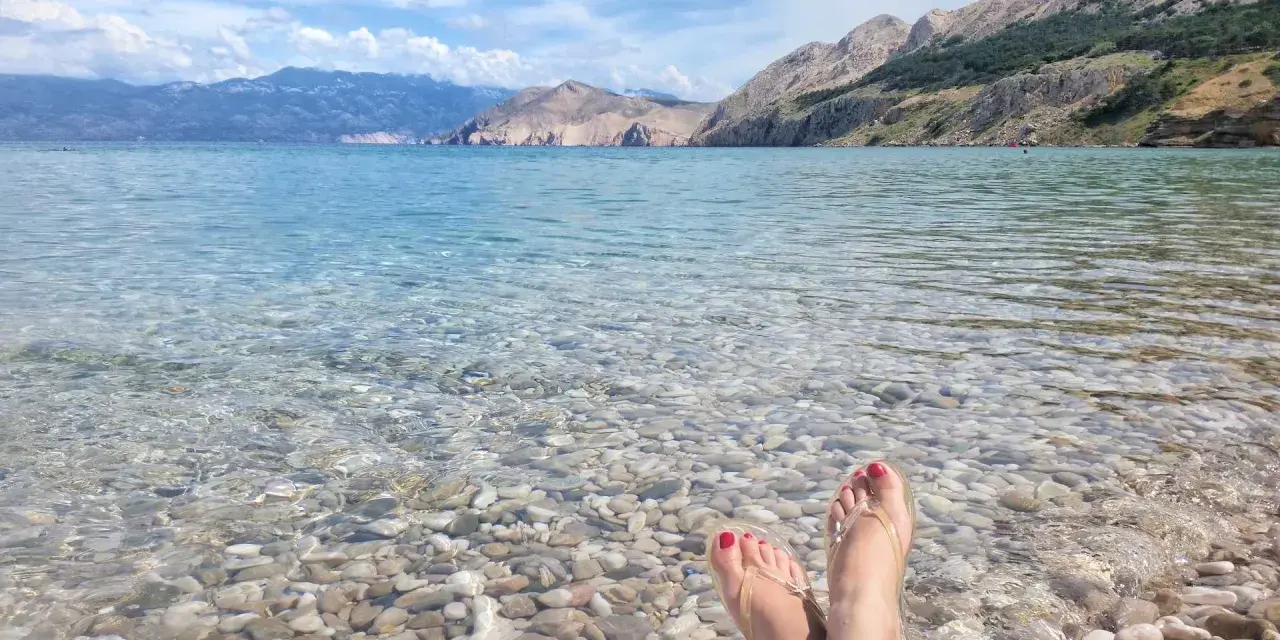 Stopy w klapkach na kamienistej plaży w **Krk National Park**. Woda krystalicznie czysta, w tle góry.
