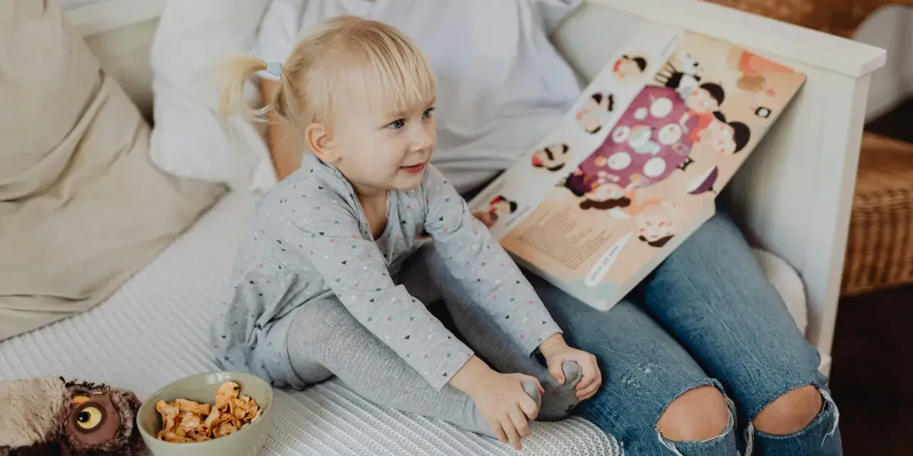 Mała dziewczynka z blond kucykami słucha bajki czytanej przez opiekunkę. Zastanawiasz się, ile zarabia opiekunka dla dziecka?