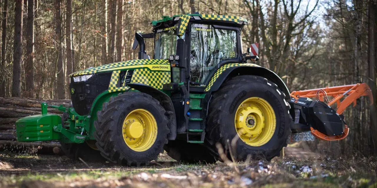 Zielony traktor John Deere z pomarańczowym mulczerem, gotowy do pracy w lesie.