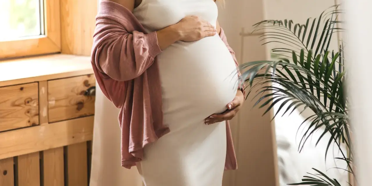 Frau in weißem Kleid und rosafarbenem Cardigan hält ihren Bauch, 34 ssw. Ein Fenster und eine Pflanze sind im Hintergrund.