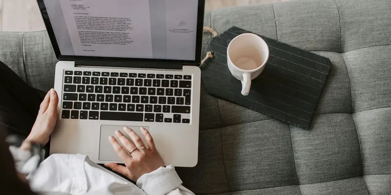 Person arbetar på laptop i soffan, en kopp står bredvid. Perfekt för dig som söker bra utbildningar på distans.