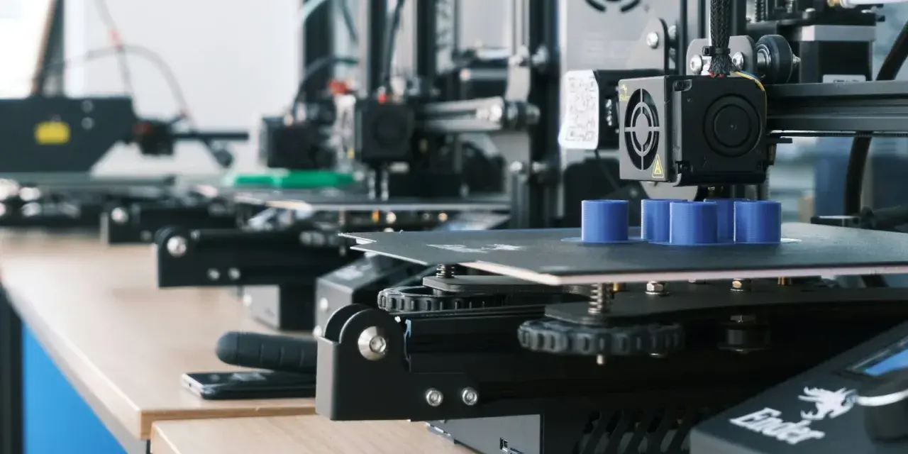 Drukarki 3D pracują, tworząc niebieskie cylindry. W tle widać kolejne maszyny, a na biurku leży smartfon, gotowy do zarządzania drukiem przez Raspberry Pi z OctoPrint.