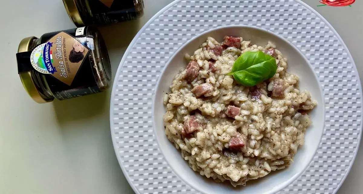 Aksamitne risotto z kawałkami kiełbasy, udekorowane listkiem bazylii. Obok stoją słoiczki z sosem truflowym, sugerujące jego obecność w daniu.