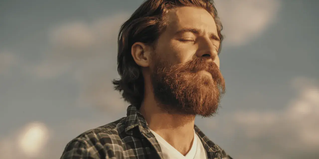 Homme à la barbe rousse, yeux fermés, profitant du soleil. Une image qui évoque le bien-être et la vitalité, peut-être liée à un test libido homme.