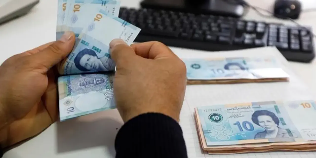 Ręce liczą banknoty 10 dinarów, tunezyjska waluta. Na stole leżą stosy pieniędzy, a w tle widać klawiaturę komputera.