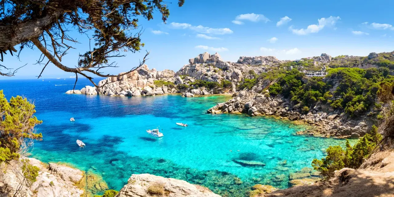 Turkusowa woda i skaliste wybrzeże Costa Smeralda na Sardynii. Kilka łodzi kołysze się na falach.