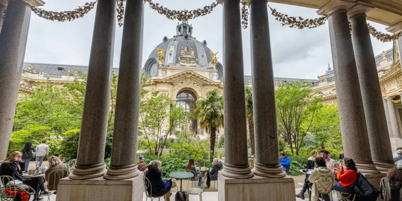Vue sur le Grand Palais depuis un portique, avec des visiteurs profitant d'un café dans le jardin. Un petit palais, un grand palais.