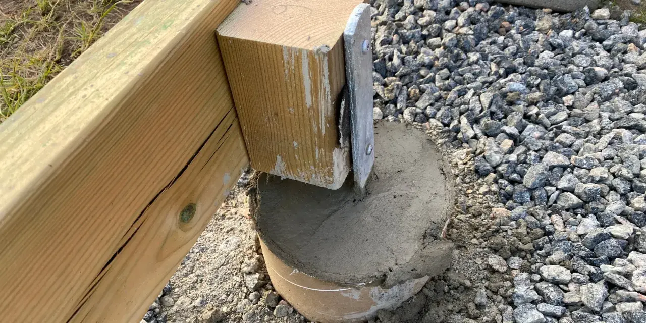 En trästolpe fästs i en betongplint, en del av arbetet med att bygga altan på plintar. Grus och jord syns runt plinten.