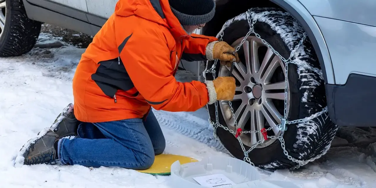 Uomo in giacca arancione monta catene da neve su ruota posteriore. Le catene da neve devono essere montate sulle ruote posteriori per la massima aderenza.