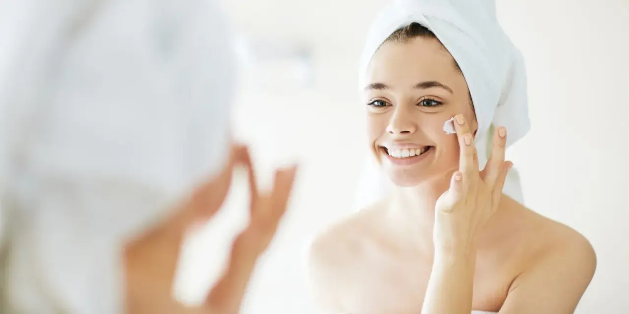 Una mujer sonriente con toalla en la cabeza se aplica crema en la cara, un paso esencial para como cuidar la piel de la cara.