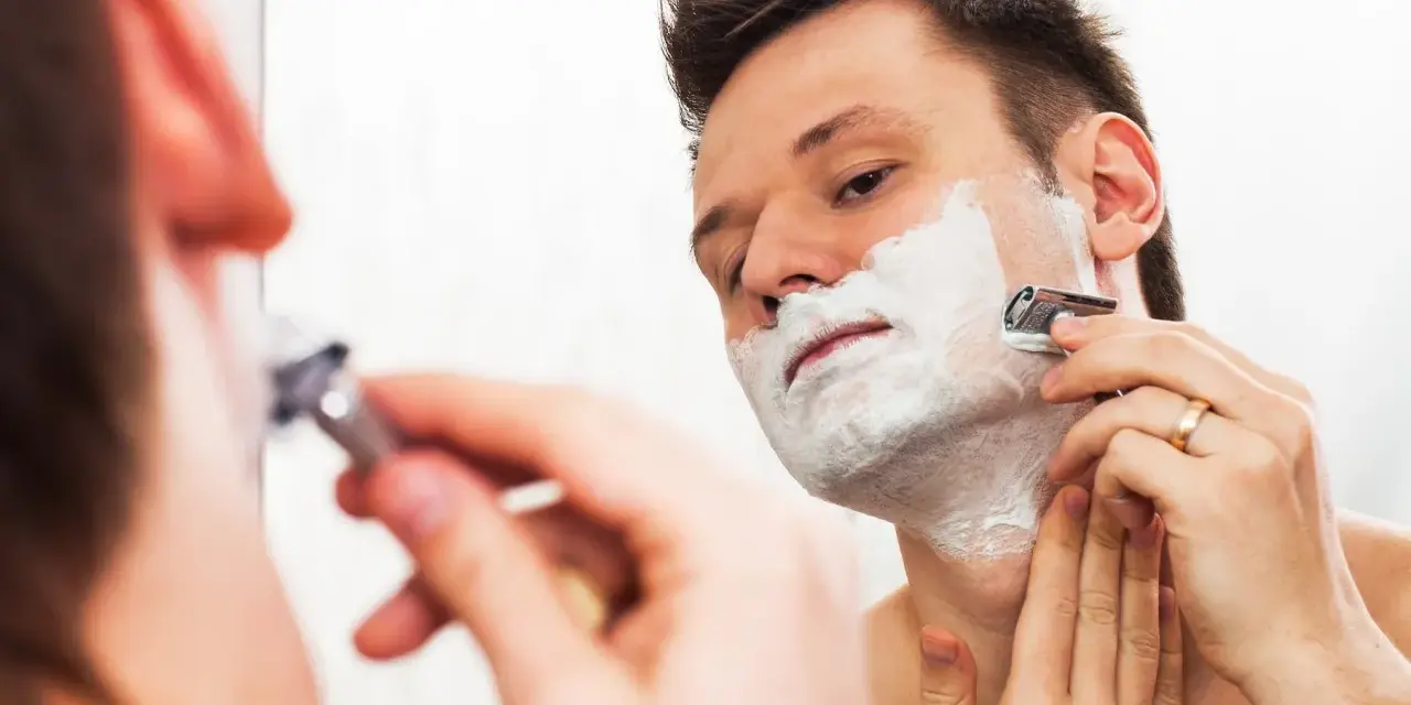 Hombre con espuma de afeitar en la cara, afeitándose con una maquinilla. ¿Es mejor afeitarse antes o después de la ducha?