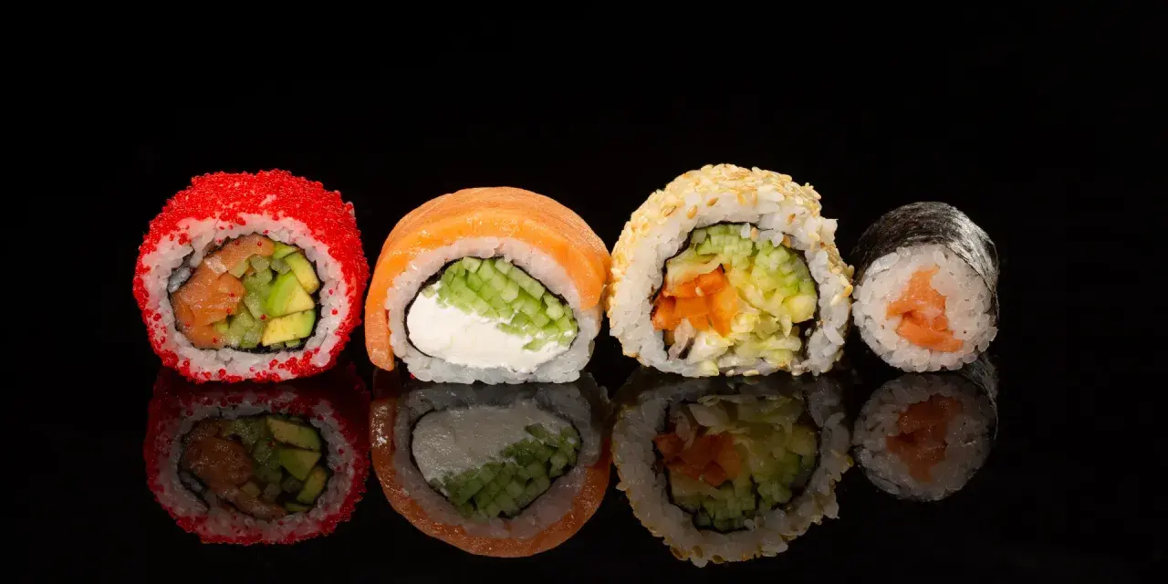 Różne rodzaje sushi: rolka z łososiem i awokado w ikrze, z łososiem i serkiem, z sezamem i warzywami, oraz klasyczne maki z łososiem.