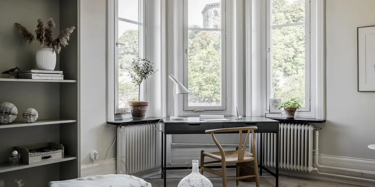Una **scrivania sotto finestra** con vista sul verde. Un angolo studio accogliente con piante, lampada e una sedia di design.