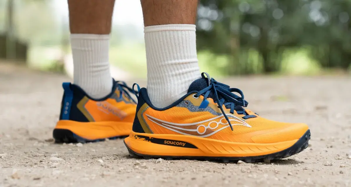 Gambe di persona che indossa scarpe da trail running Saucony arancioni e blu su un sentiero sterrato.