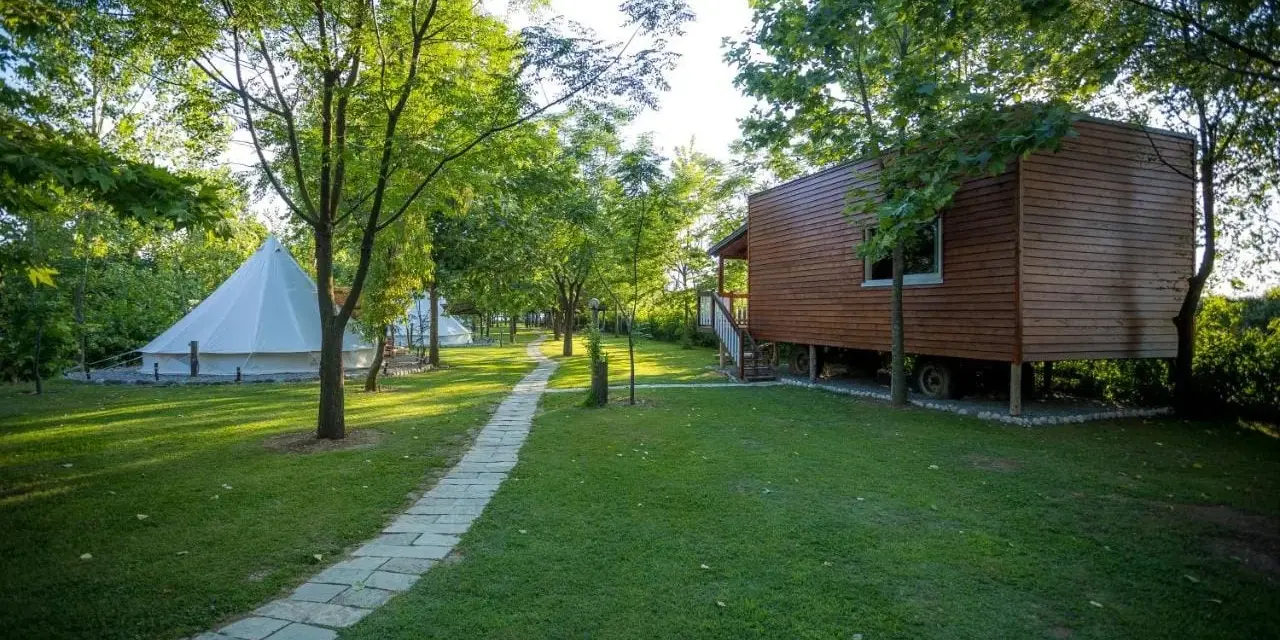 Malowniczy camping w Albanii z białymi namiotami typu bell i drewnianym domkiem na palach, otoczony zielenią.