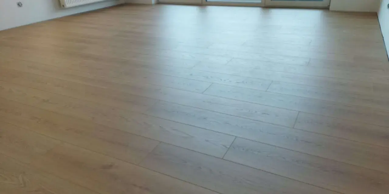 Jasne panele ułożone wzdłuż pomieszczenia, tworząc wrażenie większej przestrzeni. W tle biały grzejnik i okno balkonowe.