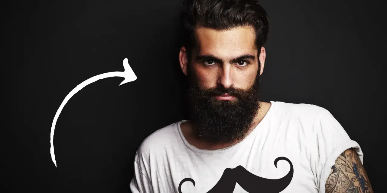 Hombre con barba tupida, quizás decidiendo dejarse barba por primera vez. Camiseta blanca con estampado de bigote.