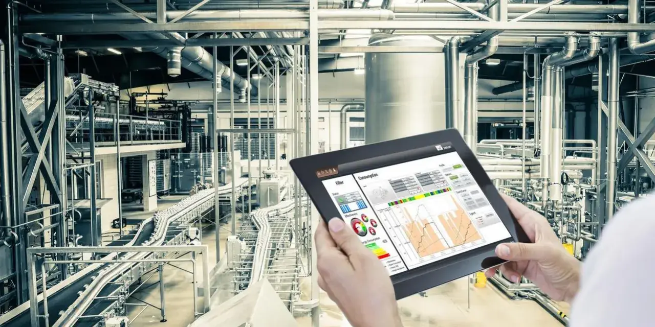 Operator kontroluje linię produkcyjną za pomocą tabletu, na którym widnieje wykres zużycia energii. W tle widać skomplikowany system rur i taśmociągów. Czujnik podczerwieni może być kluczowy dla monitorowania procesów.
