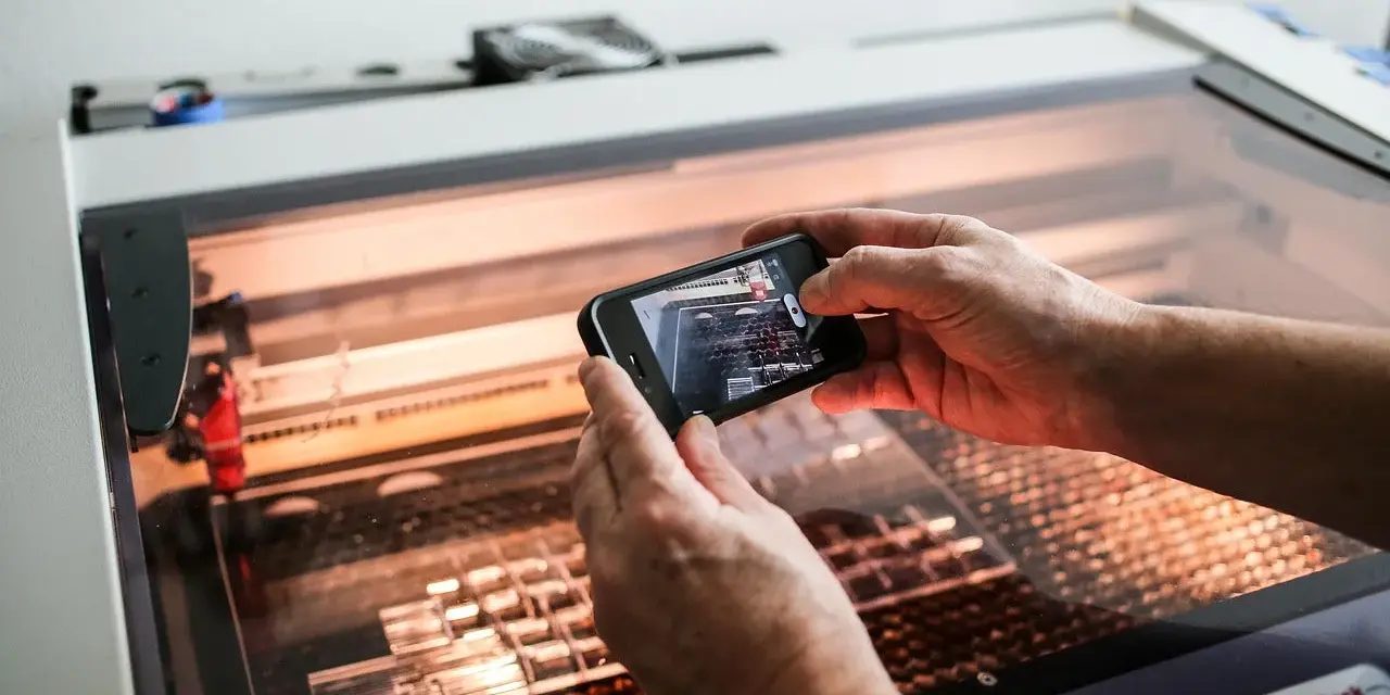 Hands hold a phone, capturing a laser cutter working on intricate acrylic projects.