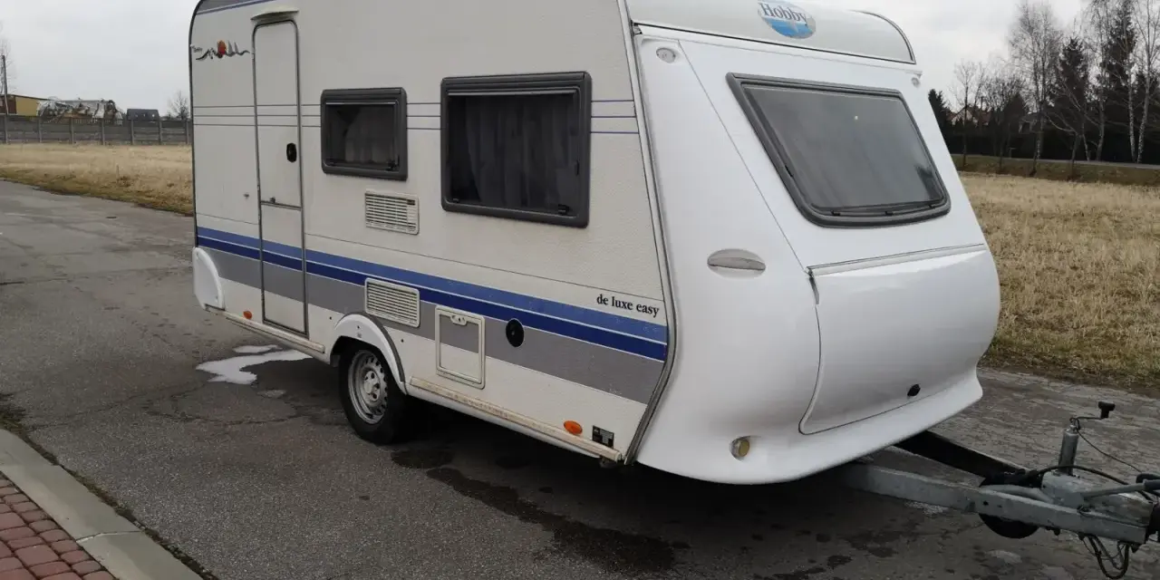 Biała przyczepa kempingowa Hobby 400 de luxe easy z niebieskimi pasami, gotowa na wakacyjne podróże.