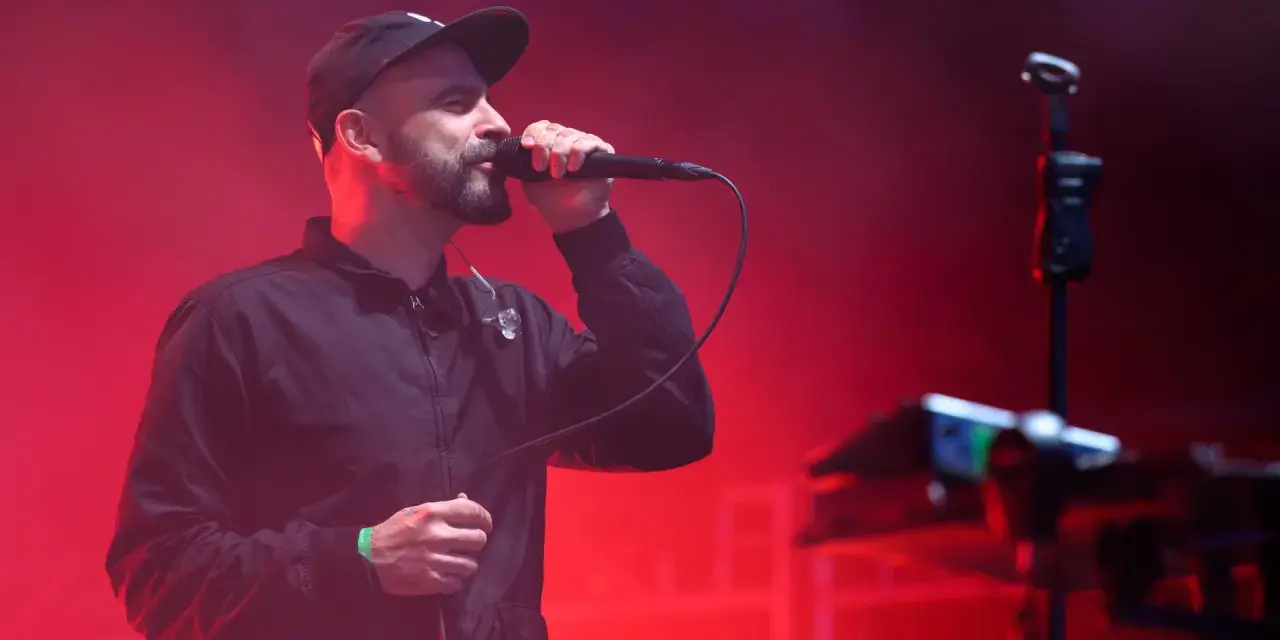 Wokalista z brodą i czapką na głowie śpiewa do mikrofonu na scenie. Czerwone światła tworzą atmosferę koncertu. Tauron Nowa Muzyka line-up.