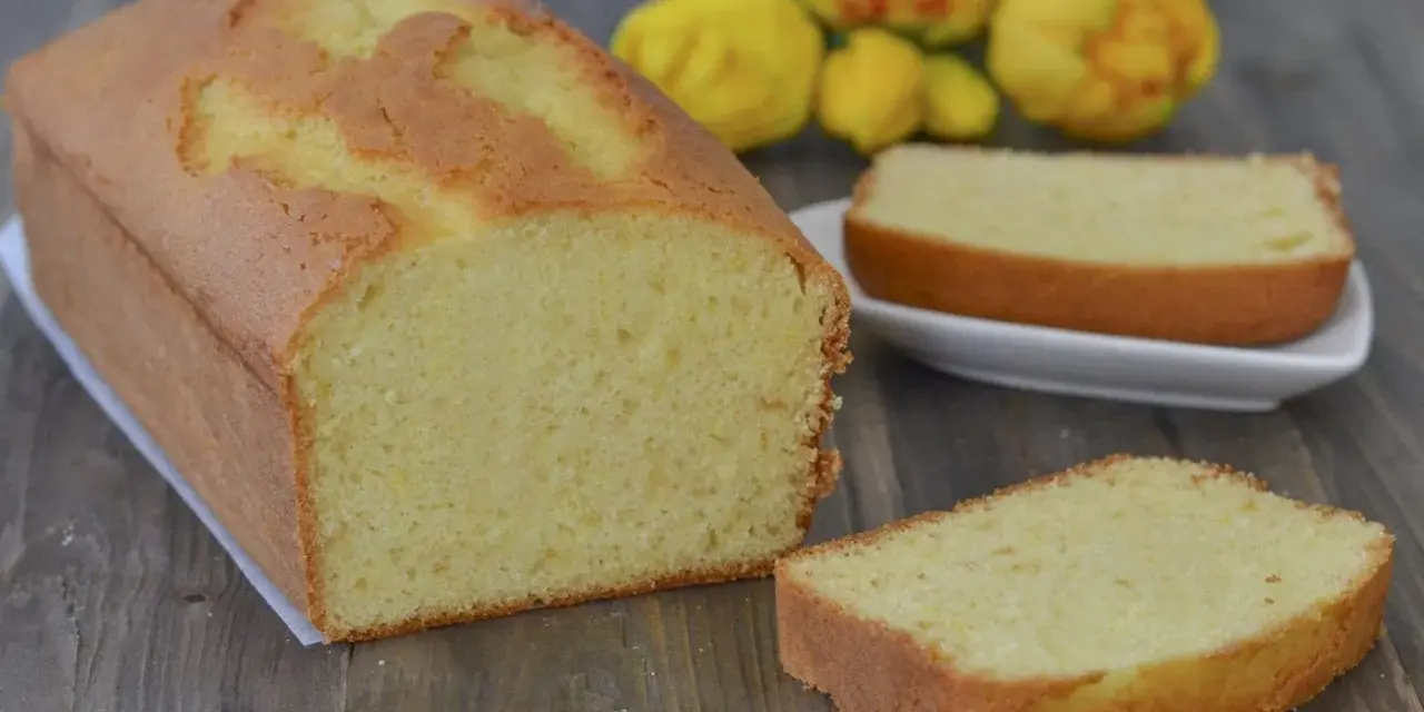 Un soffice plumcake al limone, appena sfornato, con una fetta su un piattino bianco e fiori gialli sullo sfondo.
