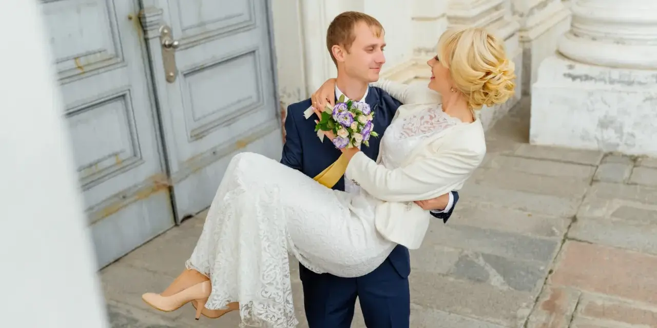 Pan młody niesie pannę młodą na rękach. Zastanawiasz się nad skutkami spóźnienia się z rezerwacją fotografa? Dlaczego 18 miesięcy to minimum?